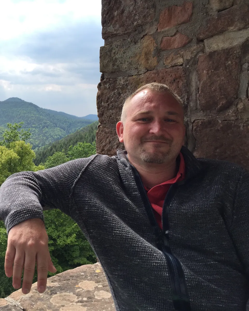 Karsten Zimmer lehnt an einer Burgmauer aus Sandstein, im Hintergrund bewaldete Hügel